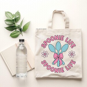 Peut inclure: Un sac fourre-tout en toile blanche avec des cuill&egrave;res roses et bleues et le texte "Spoonie Life" imprim&eacute; dessus. Le sac a un n&oelig;ud rose et deux fleurs roses de chaque c&ocirc;t&eacute; des cuill&egrave;res.