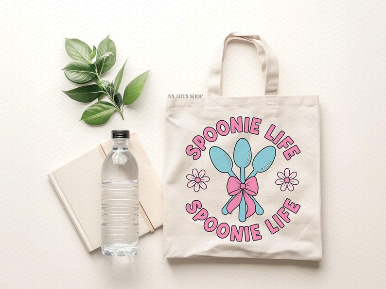 Peut inclure: Un sac fourre-tout en toile blanche avec des cuill&egrave;res roses et bleues et le texte "Spoonie Life" imprim&eacute; dessus. Le sac a un n&oelig;ud rose et deux fleurs roses de chaque c&ocirc;t&eacute; des cuill&egrave;res.