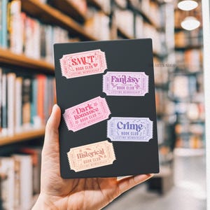Puede incluir: Un objeto rectangular negro con cinco pegatinas coloridas en forma de boleto. Las pegatinas dicen "SMUT", "Fantasy", "Dark Romance", "Crime" e "Historical" con el texto "Book Club" y "Lifetime Membership".