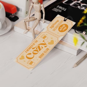May include: A bookmark with the text "A Member of the Cozy Book Club" in orange and white. The bookmark has a ticket design with coffee cup and heart illustrations. A wooden figure sits on a stack of books.