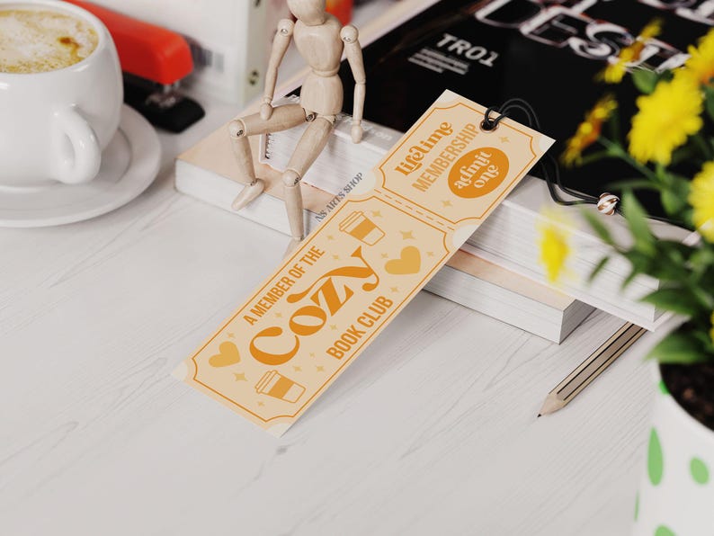 May include: A bookmark with the text "A Member of the Cozy Book Club" in orange and white. The bookmark has a ticket design with coffee cup and heart illustrations. A wooden figure sits on a stack of books.