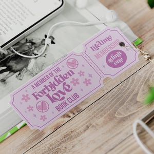 Op de afbeelding: Een lila bladwijzer met de tekst "A Member of the Forbidden Love Book Club" en "Lifetime Membership Admit One". De bladwijzer heeft een kwast en ligt naast een boek en een telefoon met oortelefoons.