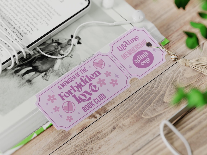 Op de afbeelding: Een lila bladwijzer met de tekst "A Member of the Forbidden Love Book Club" en "Lifetime Membership Admit One". De bladwijzer heeft een kwast en ligt naast een boek en een telefoon met oortelefoons.
