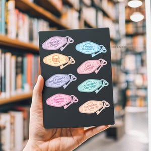 May include: A selection of colourful motel-style key tag stickers with phrases such as "Friends to Lovers" and "Arranged Marriage." Each tag features a key illustration. The stickers are held against a blurred background of bookshelves.