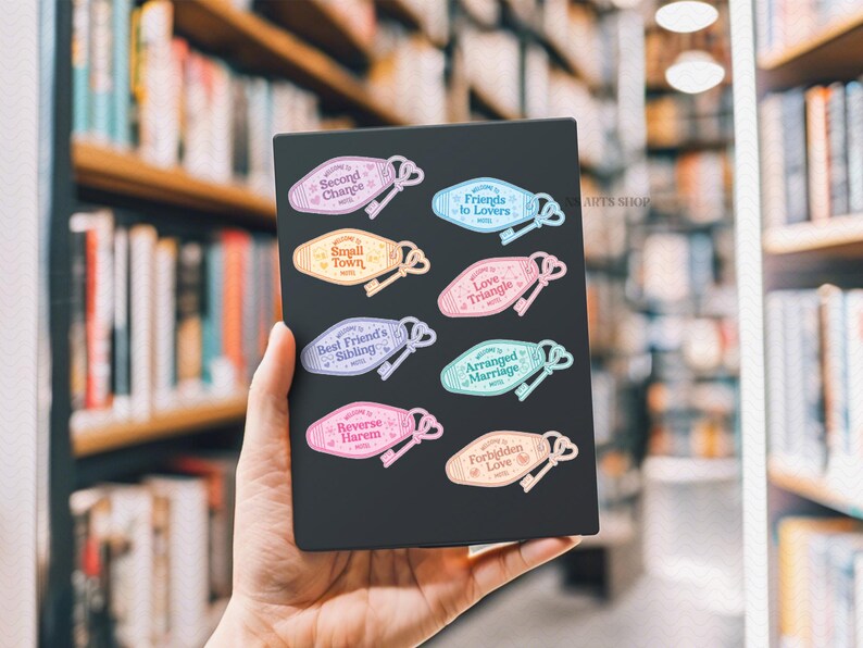 May include: A selection of colourful motel-style key tag stickers with phrases such as "Friends to Lovers" and "Arranged Marriage." Each tag features a key illustration. The stickers are held against a blurred background of bookshelves.