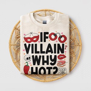 Puede incluir: Sudadera color crema con el texto "IF VILLAIN WHY HOT?" en negro. El dise&ntilde;o incluye ilustraciones rojas de una m&aacute;scara, esposas, labios, una calavera y un chile. La camiseta se muestra en una cesta tejida.