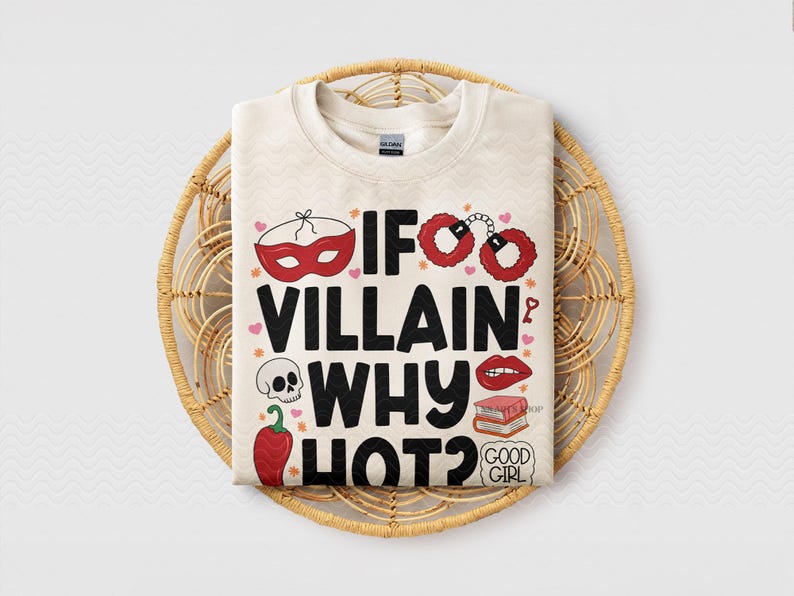Puede incluir: Sudadera color crema con el texto "IF VILLAIN WHY HOT?" en negro. El dise&ntilde;o incluye ilustraciones rojas de una m&aacute;scara, esposas, labios, una calavera y un chile. La camiseta se muestra en una cesta tejida.