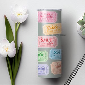 Puede incluir: Un vaso gris con dise&ntilde;os de boletos coloridos para varios clubes de lectura, incluyendo "Dark Romance", "Cozy" y "Smut". El vaso est&aacute; decorado con detalles florales y el texto "NS ARTS SHOP".