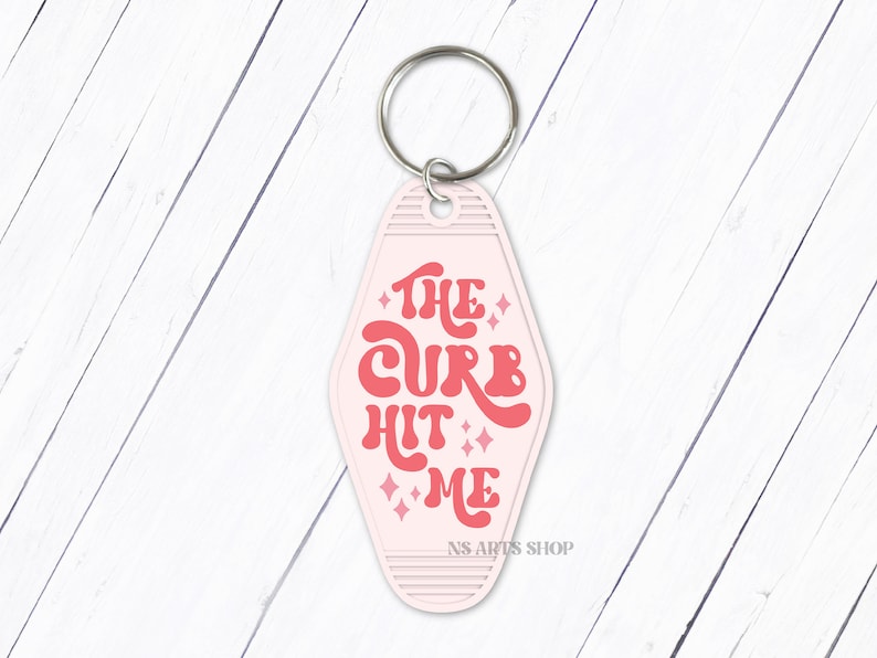 May include: A pink motel-style keychain with the phrase "THE CURB HIT ME" in red retro font. The keychain has a silver key ring and the text "NS ARTS SHOP" at the bottom.
