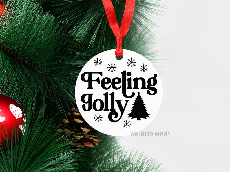 以下が含まれることがあります： 黒い文字で「Feeling Jolly」と書いてある白い丸いクリスマスオーナメント。クリスマスツリーの黒いシルエットと、黒い雪の結晶が文字の周りにあります。