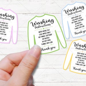 May include: A hand holding four sweater-shaped stickers with washing instructions. Each sticker has a different colored outline: pink, green, blue, and yellow. The text on each sticker reads "Washing Instructions" and laundry care guidelines.