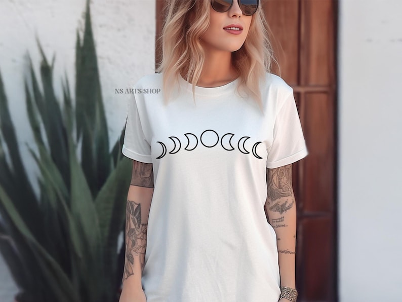 May include: White t-shirt with a black graphic of a moon phase cycle. The graphic shows the moon in different phases, from a crescent to a full moon.
