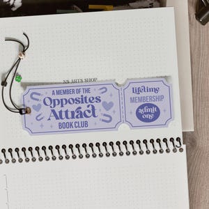 Op de afbeelding: Een lichtpaarse bladwijzer met de tekst "A Member of the Opposites Attract Book Club" en "Lifetime Membership". De bladwijzer is bevestigd aan een spiraalgebonden notitieboek met een zwart koord en een kleine groene klaver.
