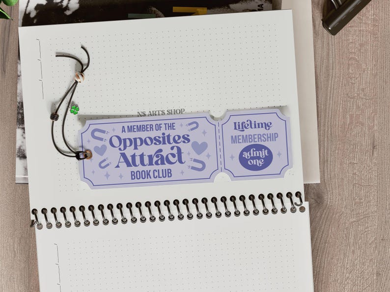 Op de afbeelding: Een lichtpaarse bladwijzer met de tekst "A Member of the Opposites Attract Book Club" en "Lifetime Membership". De bladwijzer is bevestigd aan een spiraalgebonden notitieboek met een zwart koord en een kleine groene klaver.