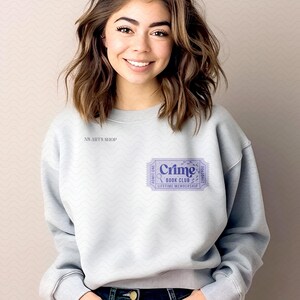 Puede incluir: Sudadera gris claro con el texto "Crime Book Club" impreso en un dise&ntilde;o de boleto morado. La sudadera tambi&eacute;n tiene el texto "NS Arts Shop" impreso en la parte superior izquierda.
