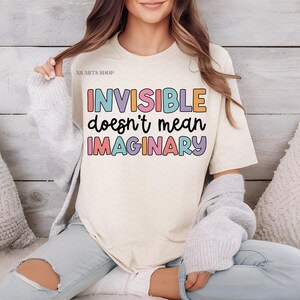 Peut inclure: Un t-shirt blanc avec le texte "INVISIBLE ne veut pas dire IMAGINAIRE" en lettres color&eacute;es. Le texte est dans une police de caract&egrave;res ludique et le t-shirt est de style d&eacute;contract&eacute;.