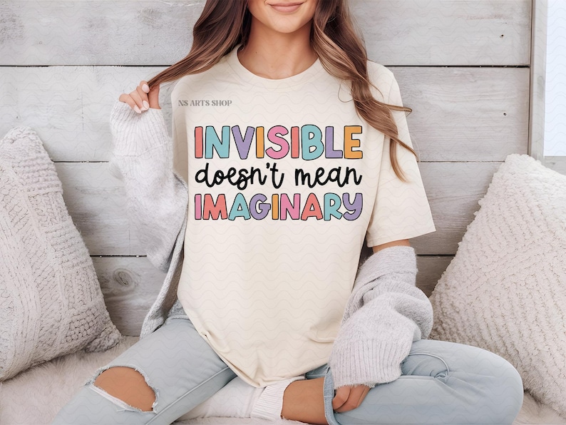 Peut inclure: Un t-shirt blanc avec le texte "INVISIBLE ne veut pas dire IMAGINAIRE" en lettres color&eacute;es. Le texte est dans une police de caract&egrave;res ludique et le t-shirt est de style d&eacute;contract&eacute;.