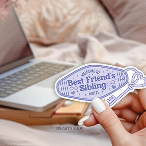 May include: A light purple sticker with the text "Welcome to Best Friend's Sibling Motel" and a key design. The sticker is held in a hand with white nail polish, with a laptop and flowers in the background.