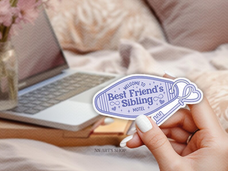 May include: A light purple sticker with the text "Welcome to Best Friend's Sibling Motel" and a key design. The sticker is held in a hand with white nail polish, with a laptop and flowers in the background.
