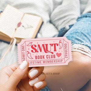 Puede incluir: Un boleto rosa con el texto "SMUT BOOK CLUB LIFETIME MEMBERSHIP" se sostiene en la mano. El boleto tiene gr&aacute;ficos de coraz&oacute;n y chile. Un libro abierto y una taza de caf&eacute; est&aacute;n en el fondo.