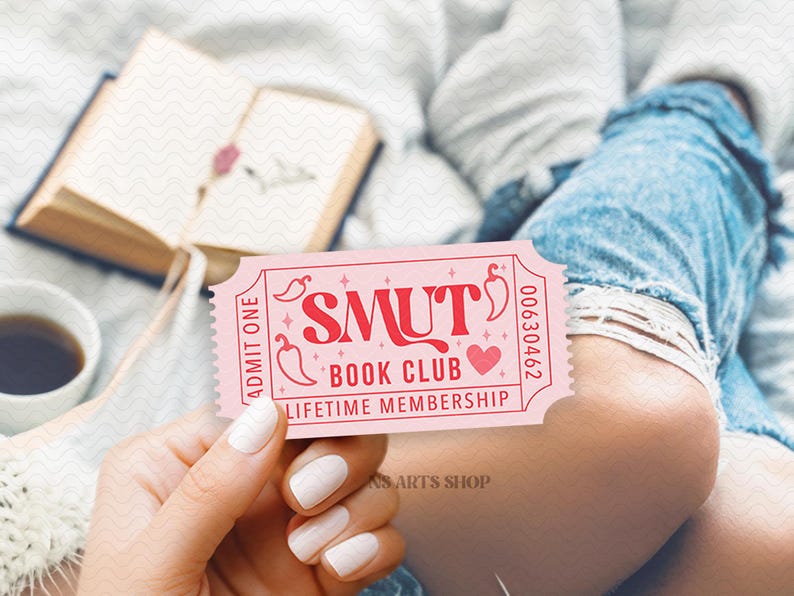 Puede incluir: Un boleto rosa con el texto "SMUT BOOK CLUB LIFETIME MEMBERSHIP" se sostiene en la mano. El boleto tiene gr&aacute;ficos de coraz&oacute;n y chile. Un libro abierto y una taza de caf&eacute; est&aacute;n en el fondo.