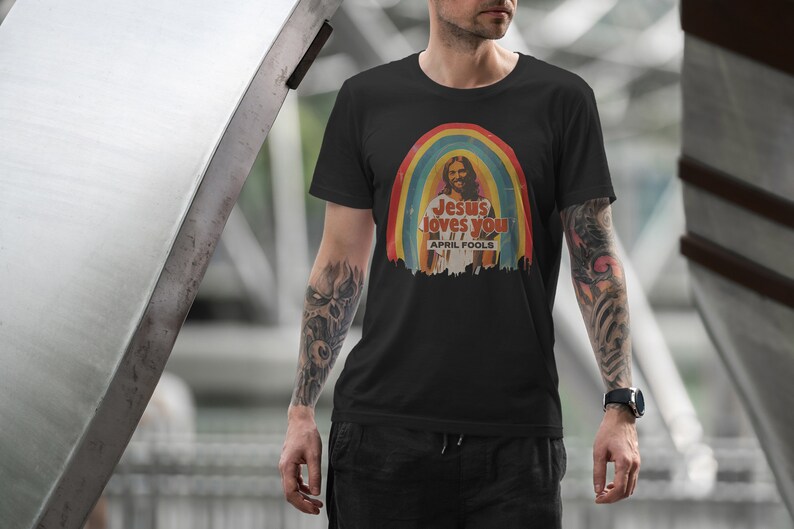 K&ouml;nnte beinhalten: Ein schwarzes T-Shirt mit einer Retro-Grafik von Jesus in einem Regenbogen mit dem Text "Jesus loves you" und "April Fools" darunter.