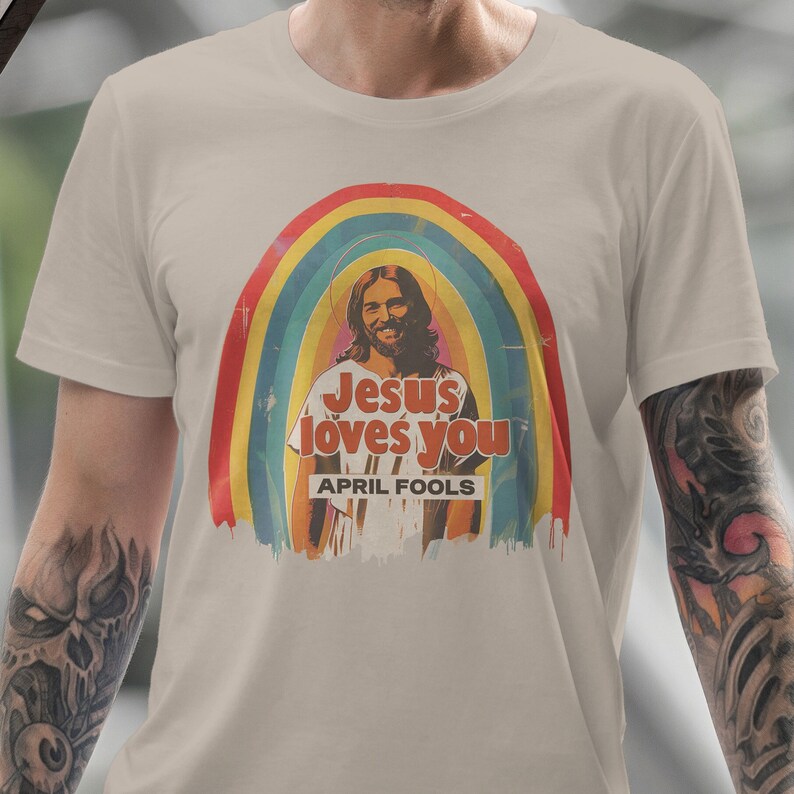K&ouml;nnte beinhalten: Ein beiges T-Shirt mit einer Retro-Grafik von Jesus mit einem Regenbogen dahinter. Der Text auf dem Shirt lautet "Jesus loves you" und "April Fools".
