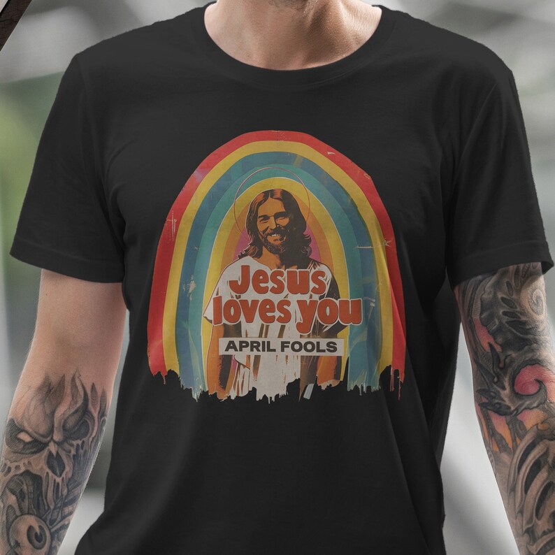 K&ouml;nnte beinhalten: Ein schwarzes T-Shirt mit einer Retro-Grafik von Jesus mit einem Regenbogen dahinter. Die Grafik lautet "Jesus liebt dich" mit dem Text "April, April" darunter.