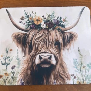 May include: A square placemat featuring a printed illustration of a brown Highland cow with long horns and shaggy fur. The cow is adorned with a colourful floral crown of yellow, purple, and white flowers, set against a light background with wildflowers.