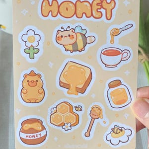 Może przedstawiać: Arkusz naklejek o tematyce miodu, zawierający różne ilustracje. Słowo "HONEY" znajduje się na górze w pomarańczowych literach. Obrazy obejmują kota-pszczołę, słoik miodu, plaster miodu i tosty z miodem.