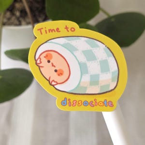 May include: A yellow sticker with the text "Time to dissociate." The sticker features a cartoon illustration of a baby wrapped in a blue and white checkered blanket. The baby has a peach-coloured face and is smiling.