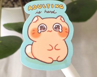 May include: A sticker featuring a cartoon cat with orange fur, rosy cheeks, and teary eyes. The cat is on a light blue background, with the words "adulting is hard" in a yellow bubble above it. The sticker is likely for use on laptops, water bottles, or notebooks.