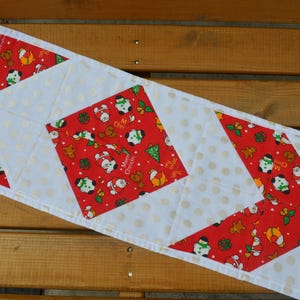 May include: A Christmas table runner with a red and white design. The runner has a repeating pattern of snowmen, Christmas trees, and the text "Merry Christmas" on a red background. The runner is approximately 30 cm wide.