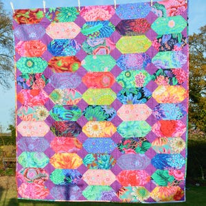 May include: A colourful quilt with a geometric pattern of hexagons and diamonds. The quilt is made of various fabrics with floral prints in shades of pink, green, blue, and yellow. The quilt is hanging on a washing line outdoors.