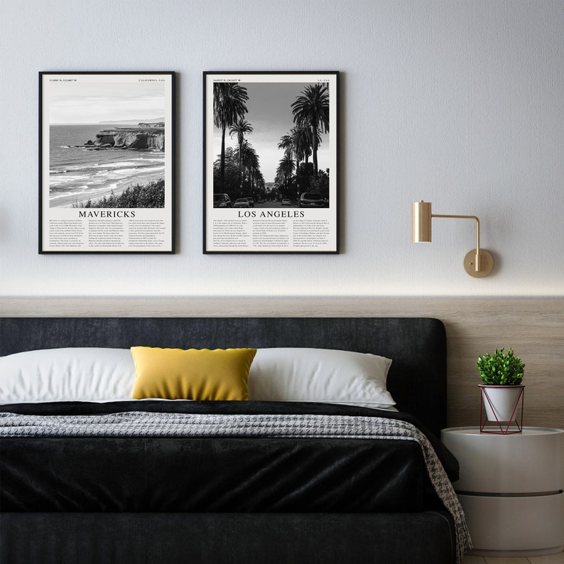 May include: Two black and white framed photographs of cityscapes. The left photo shows a beach scene with the text "MAVERICKS" below the image. The right photo shows a street lined with palm trees with the text "LOS ANGELES" below the image. The photos are hanging on a white wall above a black headboard.