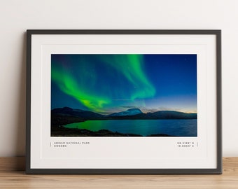 May include: Framed print featuring a vibrant display of the Northern Lights over Abisko National Park, Sweden. The image captures the green aurora borealis dancing across a dark blue sky above a serene lake and mountain range. The print is in a black frame.
