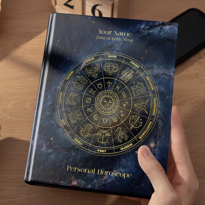 May include: A hardcover journal with a dark blue galaxy background featuring a gold foil zodiac wheel. The cover includes the text Your Name, Date of birth, Time, and Personal Horoscope. The central design displays twelve astrological signs.