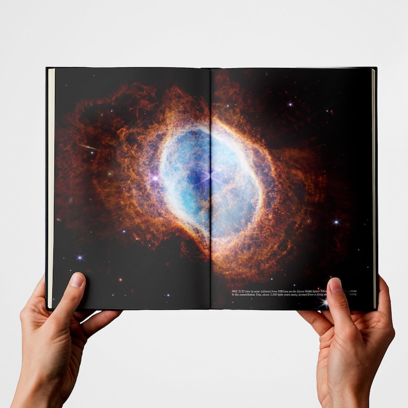 May include: An open book displaying a high-resolution space photograph of the Ring Nebula. The image features a glowing, oval-shaped cloud of orange and blue gas against a black background, with small text printed in the bottom right corner.