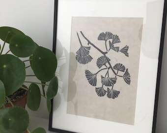 Op de afbeelding: Een zwart-wit print van een ginkgo-bladtak in een zwart frame. De print is op licht beige papier.