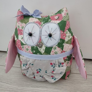 May include: A fabric owl doorstop in patchwork style, featuring green, pink, and white floral patterns. It has large white eyes with black centers, a light blue bow, and a heart charm. The wings are pink. The owl is a decorative home accessory.