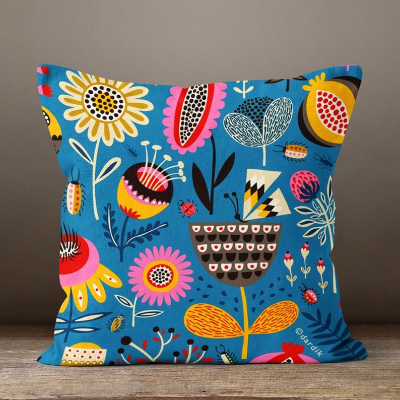 May include: Square throw pillow with a vibrant floral pattern in shades of yellow, pink, and brown on a teal background. The design includes stylized flowers, insects, and geometric shapes. The pillow is placed on a wooden surface.
