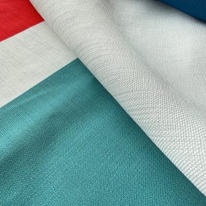 Pode incluir: Close-up de um tecido dobrado com um design geom&eacute;trico de blocos de cores. O tecido apresenta sec&ccedil;&otilde;es vermelhas, brancas, verde-azuis e azul-marinho. A textura parece ser um material tecido, com fios vis&iacute;veis e um ligeiro brilho.