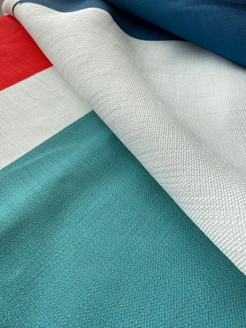 Pode incluir: Close-up de um tecido dobrado com um design geom&eacute;trico de blocos de cores. O tecido apresenta sec&ccedil;&otilde;es vermelhas, brancas, verde-azuis e azul-marinho. A textura parece ser um material tecido, com fios vis&iacute;veis e um ligeiro brilho.