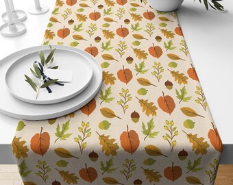 May include: A beige table runner featuring a repeating pattern of autumn leaves and acorns in orange, yellow, and green. The runner is on a white table, with white plates and candles in the background. The table runner is approximately 180 cm long.