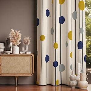 Puede incluir: Cortina color crema con un patrón repetido de líneas negras verticales y formas abstractas coloridas. Las formas son en tonos de amarillo, azul y gris. La cortina cuelga frente a una ventana. Estilo moderno de mediados de siglo.