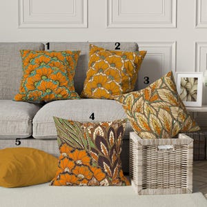 Pode incluir: Cinco almofadas decorativas com padrões florais e de folhas em tons de laranja, amarelo, castanho, verde e verde-azulado. As almofadas estão dispostas num sofá cinzento e numa cesta de vime, criando uma cena de decoração de casa acolhedora e elegante.