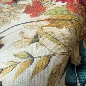 May include: Close-up of a fabric with a floral pattern. The design features large, detailed flowers in shades of red, orange, and teal, along with green and yellow leaves. The background is a light cream color, and the fabric has a textured appearance.