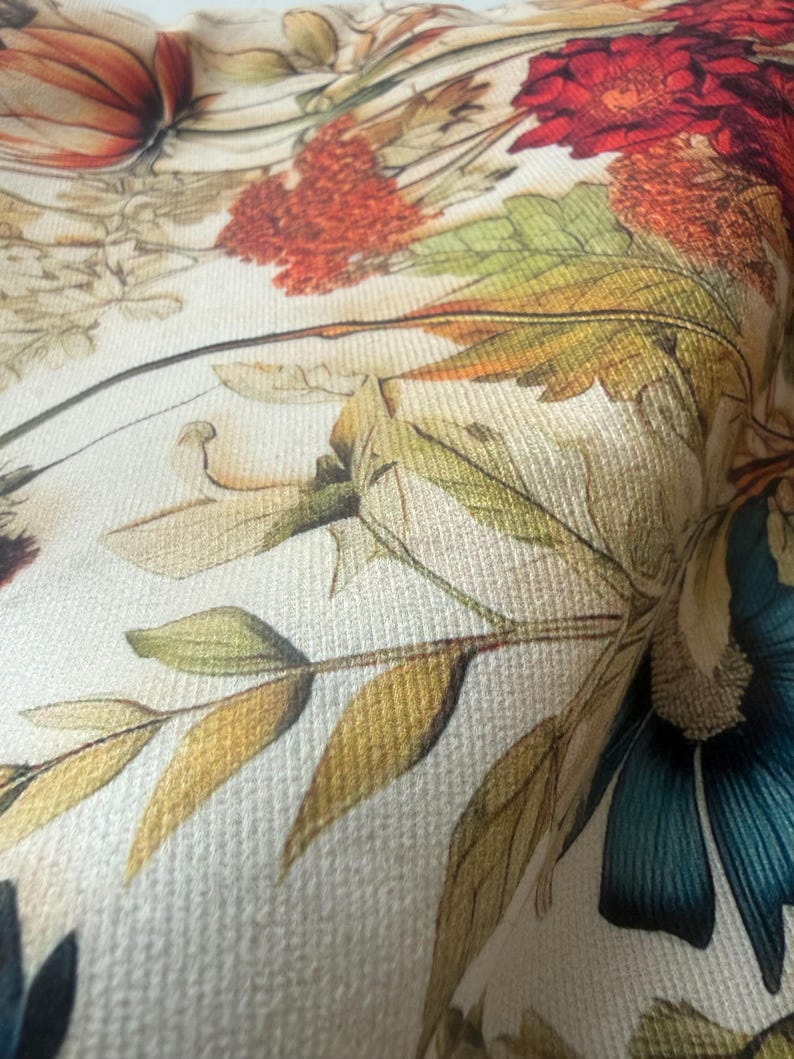May include: Close-up of a fabric with a floral pattern. The design features large, detailed flowers in shades of red, orange, and teal, along with green and yellow leaves. The background is a light cream color, and the fabric has a textured appearance.