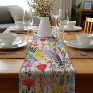 May include: A floral table runner with a cream background and a colorful wildflower design. The runner features red poppies, yellow daisies, purple lavender, and other wildflowers. The runner is set on a wooden table with white dishes and glassware.