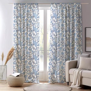 Könnte beinhalten: Ein Paar hellblau-weiß gemusterte Vorhänge vor einem Fenster. Die Vorhänge zeigen ein sich wiederholendes Muster aus stilisierten Blumen und Ranken. Der Raum umfasst auch ein beiges Sofa, einen runden Teppich und einen gewebten Hocker.